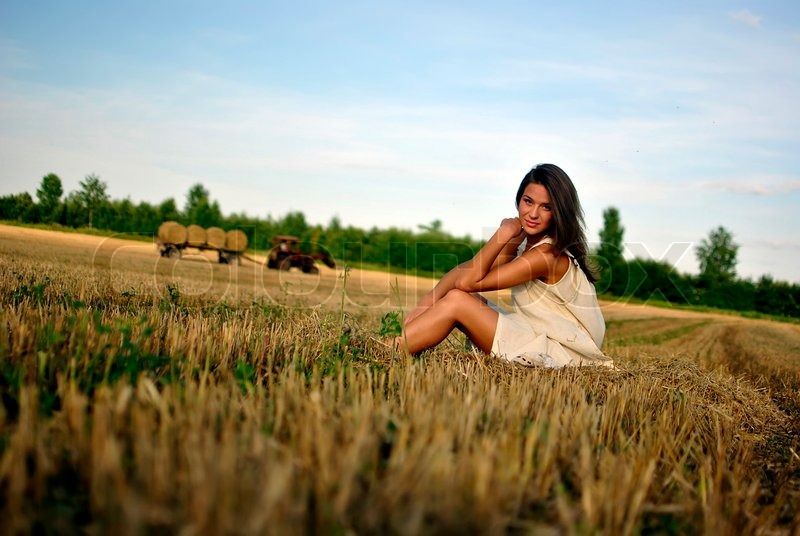 Girl in a rural clothing standing on the field, tractor on the ...
